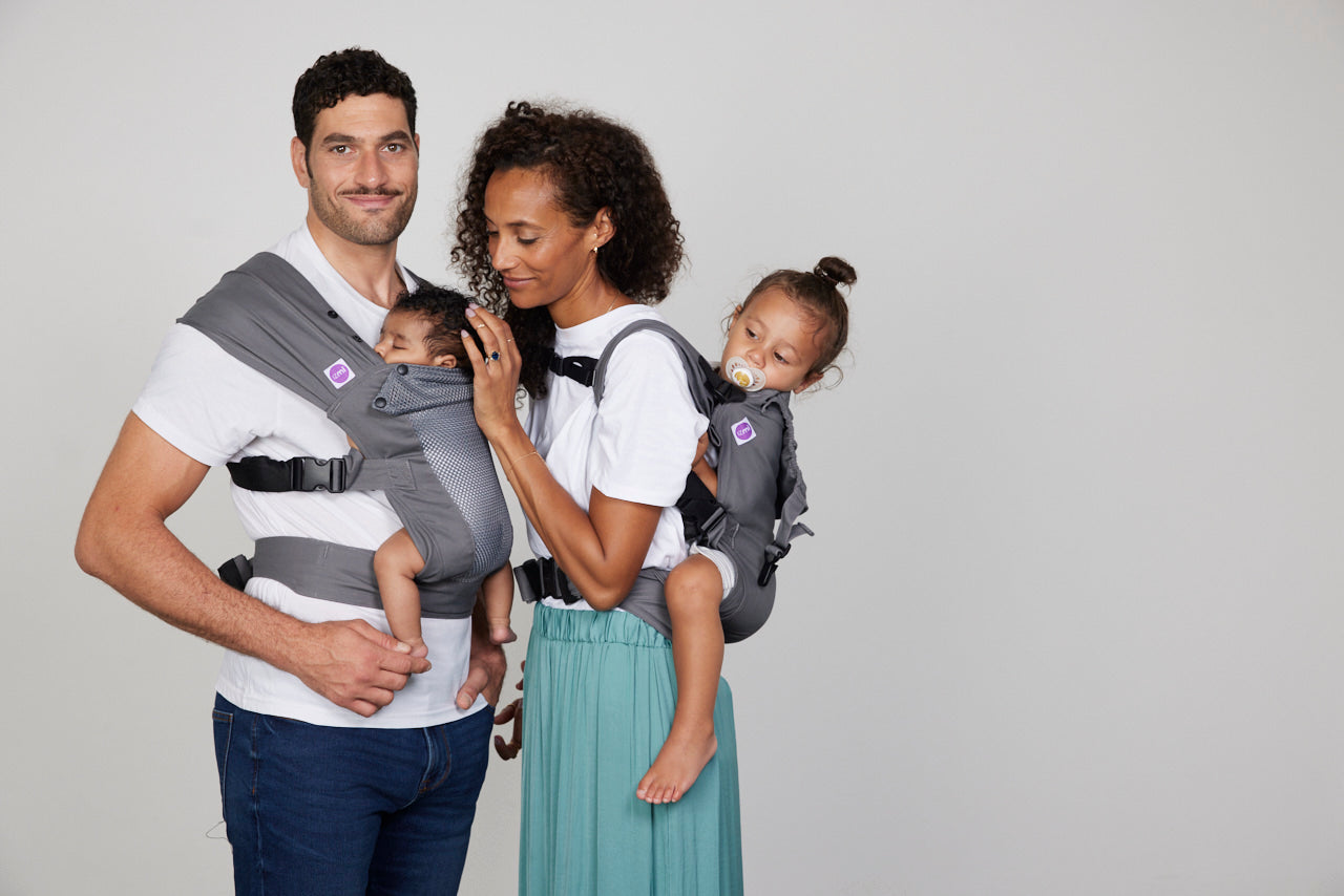Man carries baby in Izmi Baby Carrier while woman carries toddler on her back in Izmi Toddler Carrier
