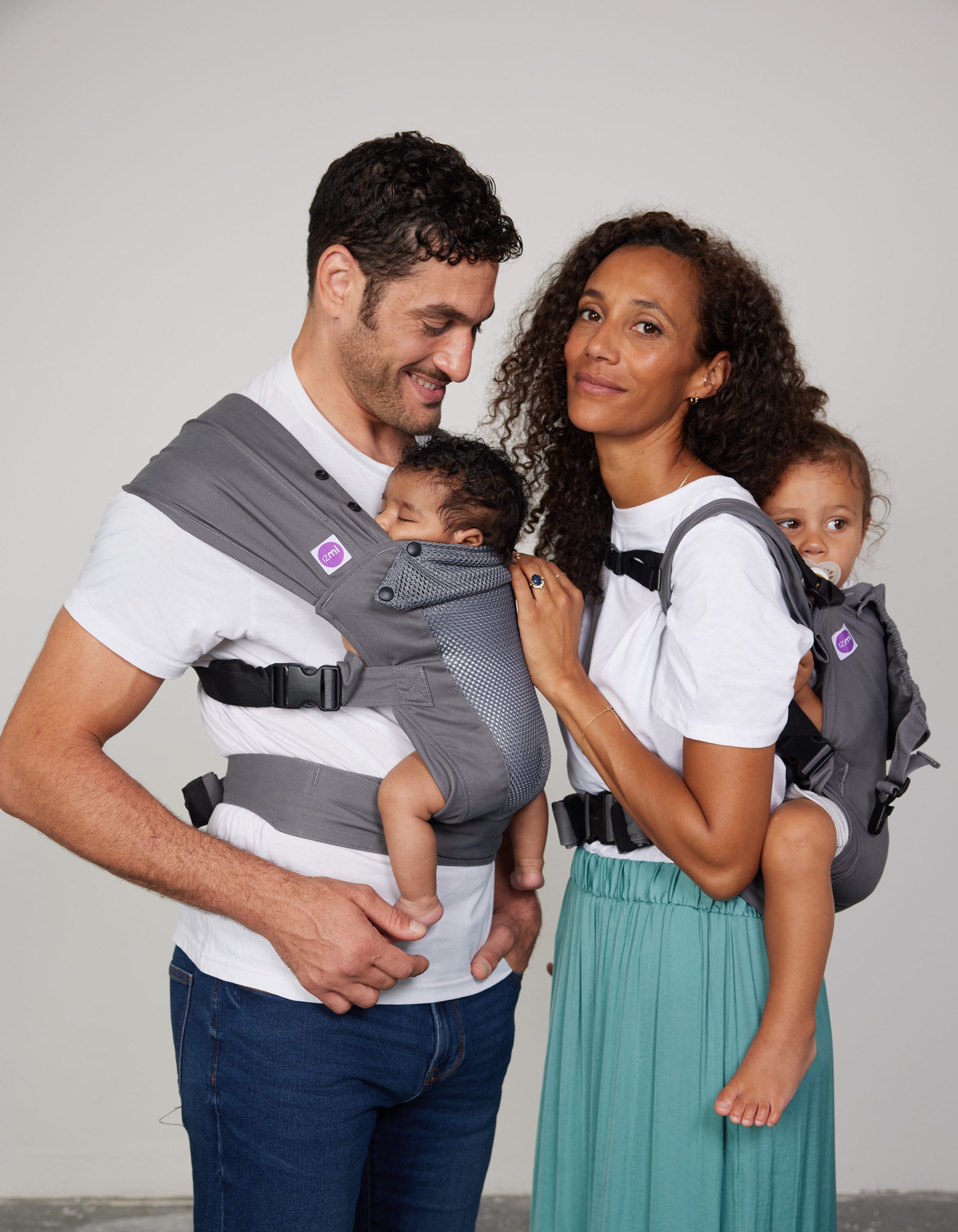 Man carries baby on his front in Izmi Breeze Baby Carrier while woman carries toddler on her back in Izmi Toddler Carrier