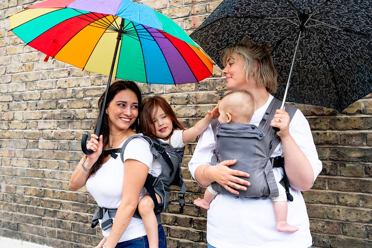 Women carry children in Izmi Toddler Carrier and Izmi Baby Carrier while holding umbrellas