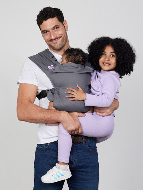 Man carries baby on his chest in Izmi Baby Carrier while holding girl on his hip