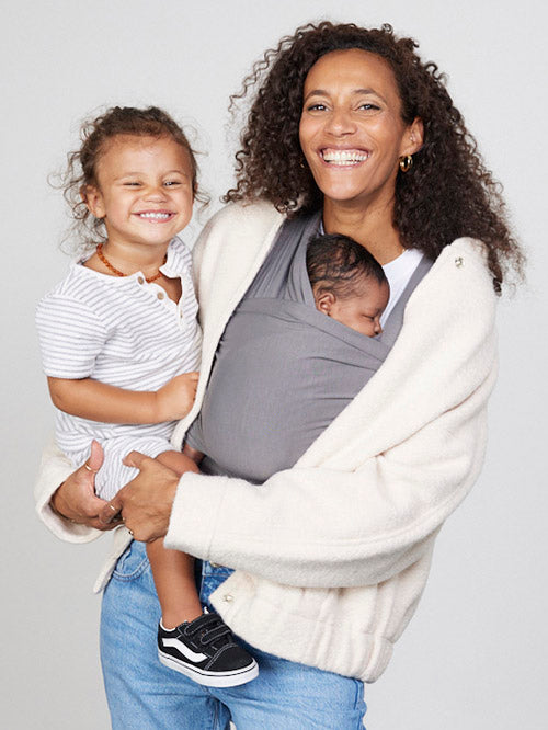 Woman carries baby facing towards her in Izmi Baby Wrap in Grey colour while holding toddler on her hip