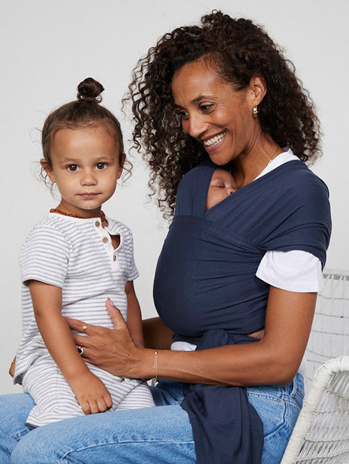 Seated woman carries baby facing towards her in Izmi Baby Wrap in Slate colour while toddler sits on her knee