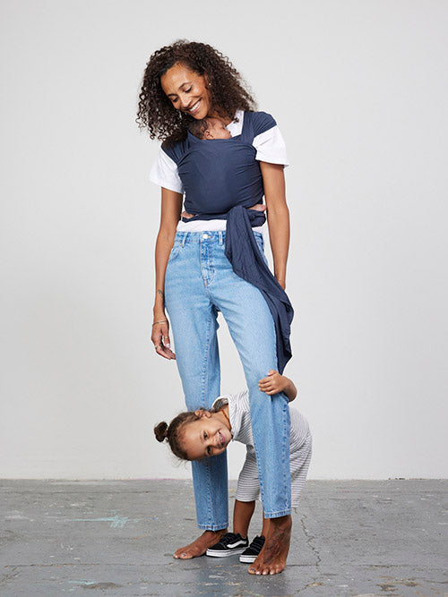 Woman carries baby facing towards her in Izmi Baby Wrap in Slate colour while toddler peeks between her knees