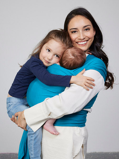 Woman carries baby facing towards her in Izmi Baby Wrap in Teal colour while also holding young girl