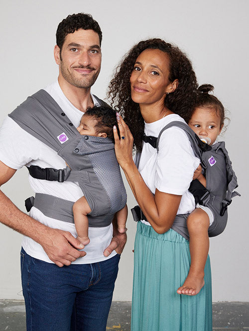 Man carries baby on his chest in Izmi Breeze Baby Carrier while woman carries toddler on her back in Izmi Toddler Carrier